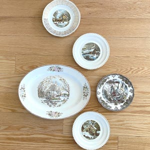 May include: A collection of five ceramic plates and one large oval platter featuring winter landscape scenes. The pieces include white backgrounds with detailed illustrations of snow-covered cabins, trees, and streams, accented by gold or patterned rims.