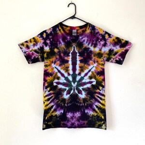 May include: A tie-dye t-shirt with a cannabis leaf design. The shirt features a vibrant mix of colors, including purple, yellow, and green, creating a psychedelic effect. The shirt is hanging on a black hanger against a white background.
