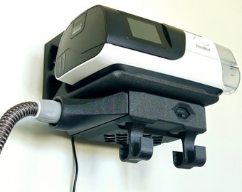 Può includere: Un apparecchio CPAP bianco e nero con un display digitale e un logo ResMed montato su un supporto a parete nero con un cavo di alimentazione e un tubo collegati.