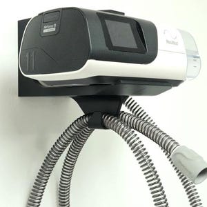 May include: A black and white AirSense 11 CPAP machine mounted on a black wall bracket. The machine has a screen and is connected to a flexible, silver hose. The ResMed logo is visible on the white portion of the machine.