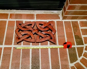 May include: Four wooden Celtic knot wall art pieces in a warm brown hue are displayed on a brick wall. A red tape measure is positioned below the art, indicating a measurement of approximately 12 inches.