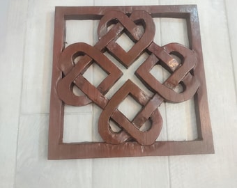 May include: A brown wooden wall art piece featuring an interwoven heart design within a square frame. The artwork has a warm, natural wood tone and a smooth finish. The design is a symbol of love and connection.