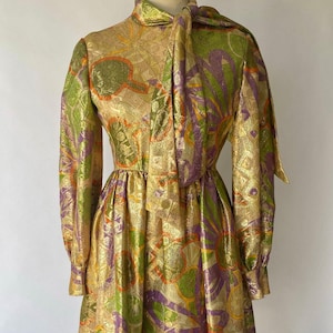 Puede incluir: Vestido dorado vintage con mangas largas y lazo en el cuello. El vestido presenta un estampado de formas verdes, moradas y naranjas. Tiene cintura entallada y falda acampanada, creando una silueta clásica.