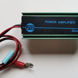 Può includere: Amplificatore di potenza nero con finiture verdi e la scritta "POWER AMPLIFIER B 35" in blu. L'amplificatore ha un logo ZG e la scritta "Zelagi-Italy". Fili rossi e neri con un connettore blu e rosso.