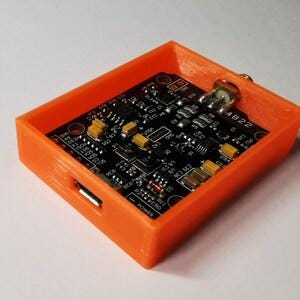 May include: A rectangular orange plastic enclosure housing a black electronic circuit board. The board features various small components, including yellow capacitors, integrated circuits, and a metallic port, with the text 4822 visible on the surface.