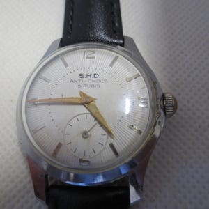 Puede incluir: Un reloj de pulsera plateado con una esfera blanca y una correa de cuero negra. La esfera del reloj tiene marcadores de hora romanos negros, una aguja de segundos dorada y el texto "S.H.D. ANTI-CHOC 15 RUBIS".