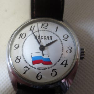 May include: A silver wristwatch with a white face and black hands. The face has black Roman numeral hour markers and a small Russian flag graphic. The watch face also has the word "Россия" printed in black.