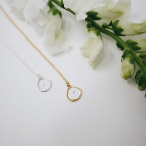 May include: Two delicate necklaces with small, round pendants. The pendants are white with a black dot in the center. One necklace has a gold chain and the other has a silver chain. The necklaces are displayed on a white surface with white flowers.