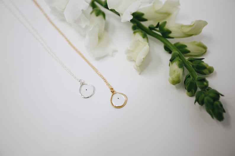 May include: Two delicate necklaces with small, round pendants. The pendants are white with a black dot in the center. One necklace has a gold chain and the other has a silver chain. The necklaces are displayed on a white surface with white flowers.