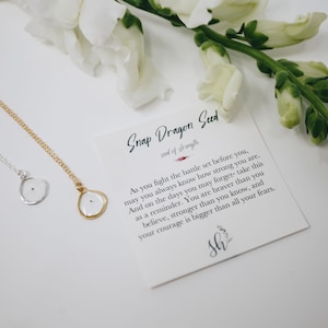 Peut inclure: Un collier en or et en argent avec un petit pendentif transparent contenant une seule graine noire. Le pendentif a la forme d'une fleur. Le collier est livré avec une carte qui dit "Snap Dragon Seed, seed of strength" et comprend une citation sur le courage et la force.