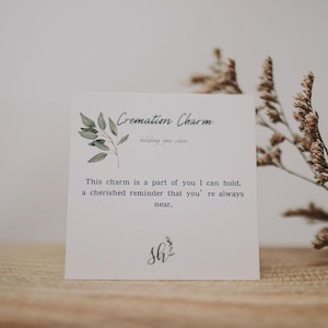 K&ouml;nnte beinhalten: Eine wei&szlig;e Karte mit gr&uuml;nen Aquarellbl&auml;ttern und dem Text "Cremation Charm" und "This charm is a part of you I can hold, a cherished reminder that you're always near." Die Karte enth&auml;lt auch den Text "Sh" in Kursivschrift.