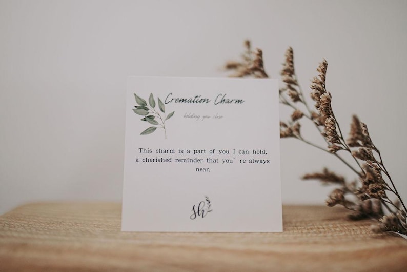 K&ouml;nnte beinhalten: Eine wei&szlig;e Karte mit gr&uuml;nen Aquarellbl&auml;ttern und dem Text "Cremation Charm" und "This charm is a part of you I can hold, a cherished reminder that you're always near." Die Karte enth&auml;lt auch den Text "Sh" in Kursivschrift.