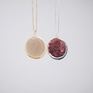 May include: Two pendant necklaces with round, textured stones. One has a gold-tone chain and bezel, the other a silver-tone chain and bezel. The stone in the gold necklace is a light beige color, while the other is a deep red.