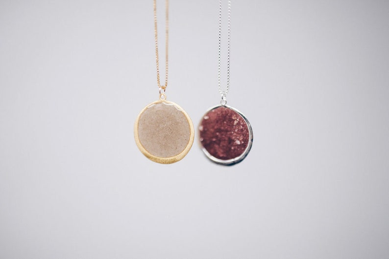 May include: Two pendant necklaces with round, textured stones. One has a gold-tone chain and bezel, the other a silver-tone chain and bezel. The stone in the gold necklace is a light beige color, while the other is a deep red.