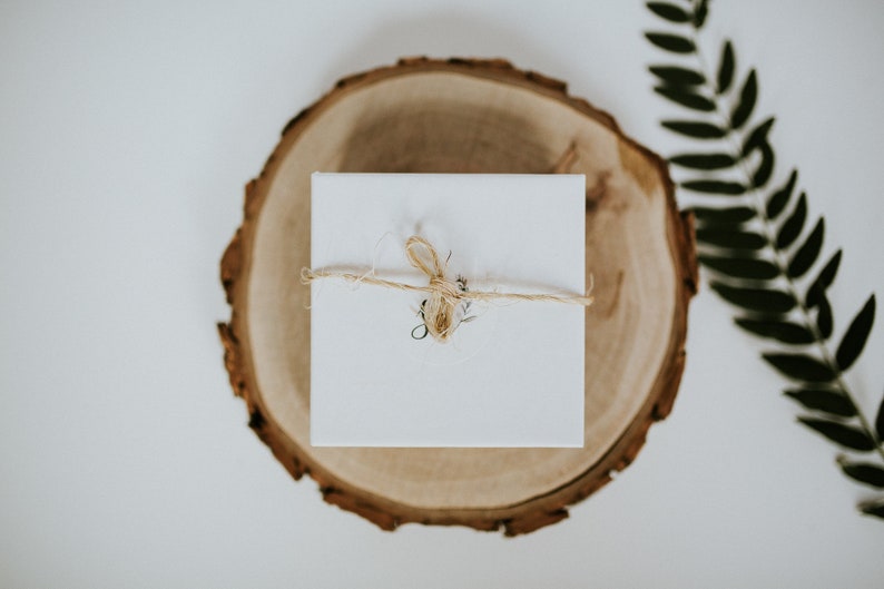 May include: A white gift box tied with twine and a small sprig of greenery, sitting on a wooden slice.