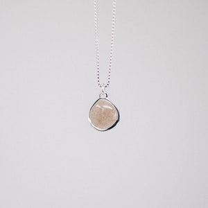 May include: A silver necklace with a teardrop-shaped pendant. The pendant has a light brown, sandy appearance and is encased in a silver bezel. The chain is a delicate silver chain. The background is a plain white.