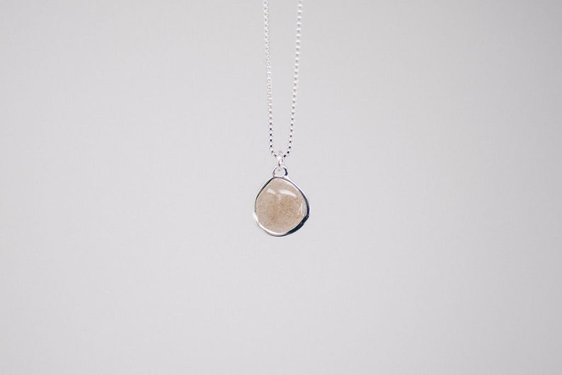 May include: A silver necklace with a teardrop-shaped pendant. The pendant has a light brown, sandy appearance and is encased in a silver bezel. The chain is a delicate silver chain. The background is a plain white.