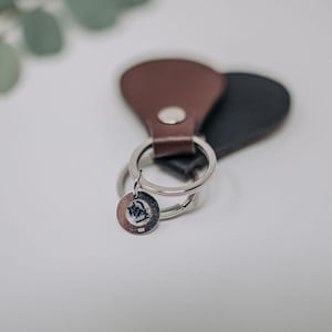 K&ouml;nnte beinhalten: Ein brauner Leder-Schl&uuml;sselanh&auml;nger mit einem silbernen Schl&uuml;sselring und einem kleinen silbernen Anh&auml;nger. Der Anh&auml;nger hat ein Design.