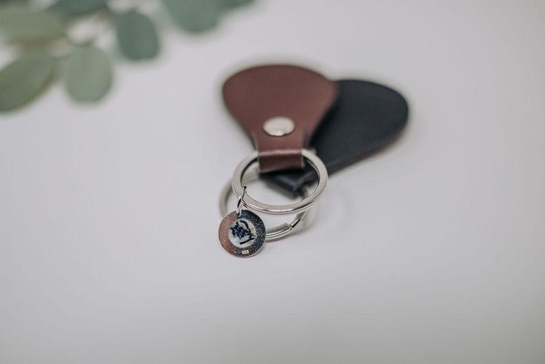 K&ouml;nnte beinhalten: Ein brauner Leder-Schl&uuml;sselanh&auml;nger mit einem silbernen Schl&uuml;sselring und einem kleinen silbernen Anh&auml;nger. Der Anh&auml;nger hat ein Design.