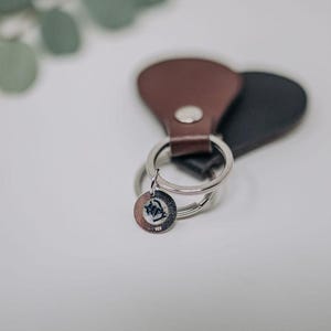 K&ouml;nnte beinhalten: Ein brauner und schwarzer Leder-Schl&uuml;sselanh&auml;nger mit einem silbernen Schl&uuml;sselring und einem kleinen silbernen Anh&auml;nger. Der Anh&auml;nger hat ein Design.