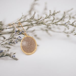 May include: A gold-colored necklace with a circular pendant. The pendant has a light brown, speckled center and a gold-colored border. The necklace is displayed on a white surface with dried flowers.