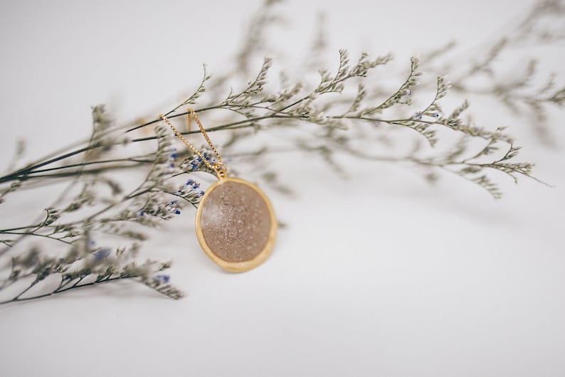 May include: A gold-colored necklace with a circular pendant. The pendant has a light brown, speckled center and a gold-colored border. The necklace is displayed on a white surface with dried flowers.