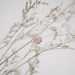 May include: A silver necklace with a pendant featuring a light brown, irregularly shaped stone. The pendant is encased in a silver bezel and hangs from a delicate chain. The background includes soft, blurred white flowers and stems.