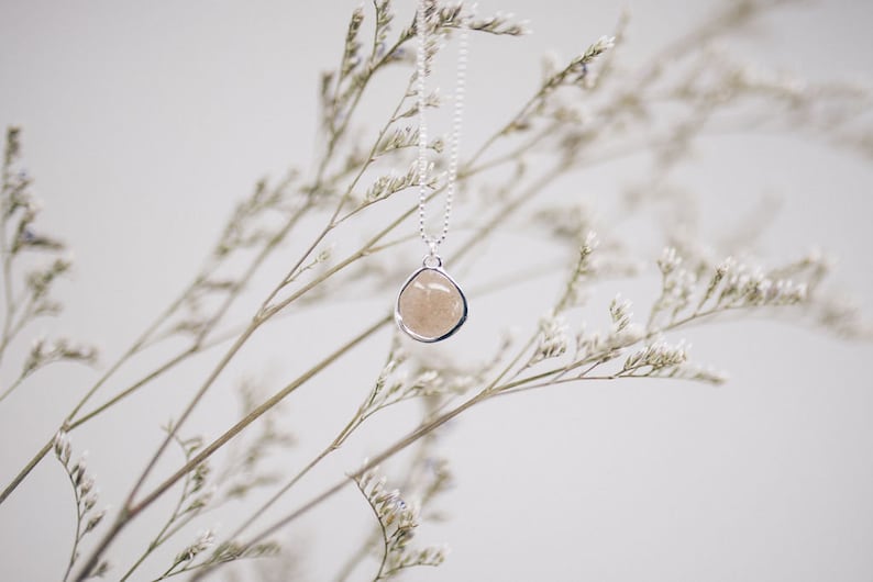 May include: A silver necklace with a pendant featuring a light brown, irregularly shaped stone. The pendant is encased in a silver bezel and hangs from a delicate chain. The background includes soft, blurred white flowers and stems.