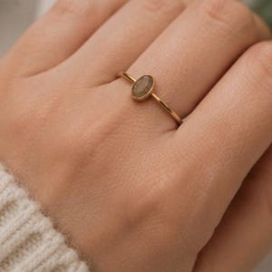 May include: A thin gold band ring featuring a small, oval-shaped, translucent taupe stone set in a simple metal bezel. The ring has a delicate, slender profile and a polished finish, showcasing a neutral-toned gemstone centered on the band.