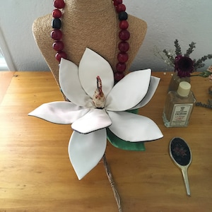 May include: A white magnolia flower with a green stem and leaves, a red and black beaded necklace on a straw-coloured mannequin, a clear glass bottle with a label that reads "25 PRINCE'S LIP STIX", and a black and red hairbrush on a wooden surface.