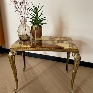 May include: A rectangular side table with a polished stone mosaic top featuring shades of brown, cream, and amber. The table has four ornate, curved gold-toned metal legs with intricate detailing and a decorative trim along the edge of the tabletop.