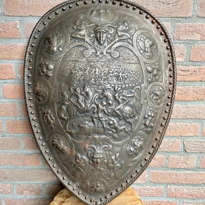 Op de afbeelding: Een decoratief metalen schild met een ingewikkeld reliëf van een veldslag met ruiters. De rand is versierd met een touwachtig patroon en klinknagels, terwijl de bovenste en onderste delen gedetailleerde menselijke gezichten tonen.