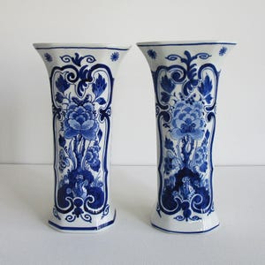 Può includere: Due vasi bianchi con disegni floreali blu. I vasi hanno una parte superiore svasata e una base rettangolare. Il design floreale blu comprende fiori, foglie e volute decorative. I vasi sono affiancati su uno sfondo bianco.