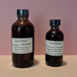 Op de afbeelding: Twee amberkleurige glazen flessen met zwarte doppen die vanille-extract bevatten. Elke fles heeft een wit etiket met de tekst Vanilla Extract, Date Bottled: 06/30/2025, Ingredients: Vodka and Organic Madagascar Vanilla Beans, Shake well and store in cool dark place.