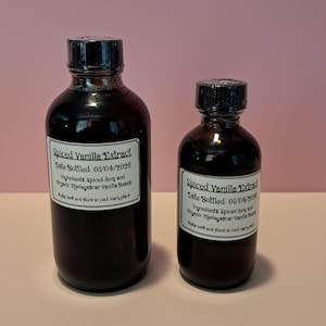 Op de afbeelding: Twee donkerbruine glazen flessen met zwarte schroefdoppen, elk voorzien van een wit rechthoekig etiket. De etiketten vermelden: Spiced Vanilla Extract, Date Bottled: 01/04/2026, Ingredients: Spiced Rum and Organic Madagascar Vanilla Beans, Shake well and store in cool, dark place.