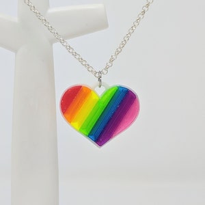 Puede incluir: Un collar de cadena de plata con un colgante de corazón arcoíris. El corazón está hecho de plástico transparente con rayas arcoíris.