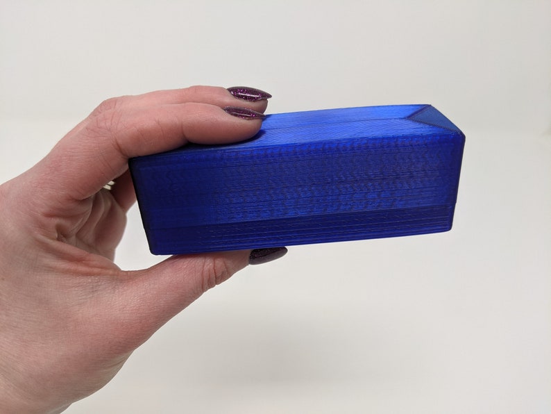 May include: A small, rectangular box made of blue plastic. The box has a smooth, glossy finish and is slightly angled on the top and bottom.