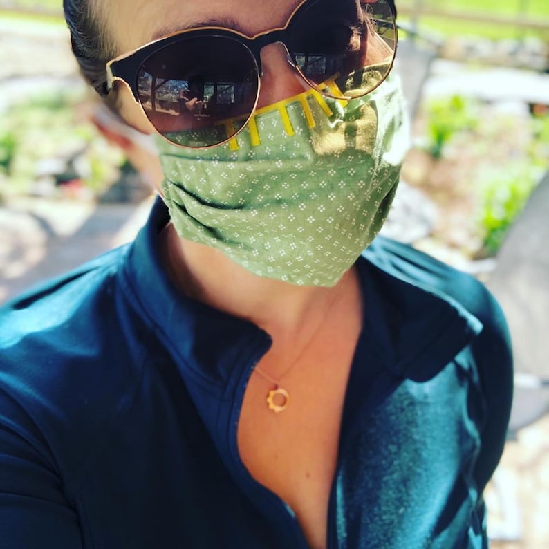 May include: A person wearing a green and white patterned face mask with yellow accents and dark sunglasses. The mask is folded and has a small white pattern. The person is wearing a dark blue jacket.