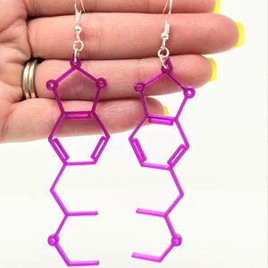 May include: A pair of purple earrings shaped like a chemical structure. The earrings are made of a 3D printed material and have silver hooks.