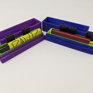 May include: Two 3D printed cases for vape pens. One case is purple with a yellow vape pen that says "D.A.L.I.Z." The other case is blue with a red vape pen.