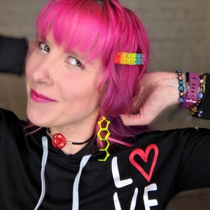 May include: A person with pink hair wearing a black sweatshirt with the word "LOVE" written in white with a red heart. They are wearing a black choker with a red geometric design and yellow geometric earrings. They are also wearing a rainbow beaded hair clip and several bracelets on their wrist.
