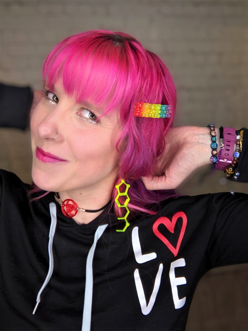May include: A person with pink hair wearing a black sweatshirt with the word "LOVE" written in white with a red heart. They are wearing a black choker with a red geometric design and yellow geometric earrings. They are also wearing a rainbow beaded hair clip and several bracelets on their wrist.