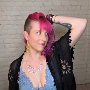 May include: A woman with pink hair and a shaved head is wearing a black sheer top with floral embroidery, a blue lace bralette, a geometric necklace, and yellow geometric earrings.
