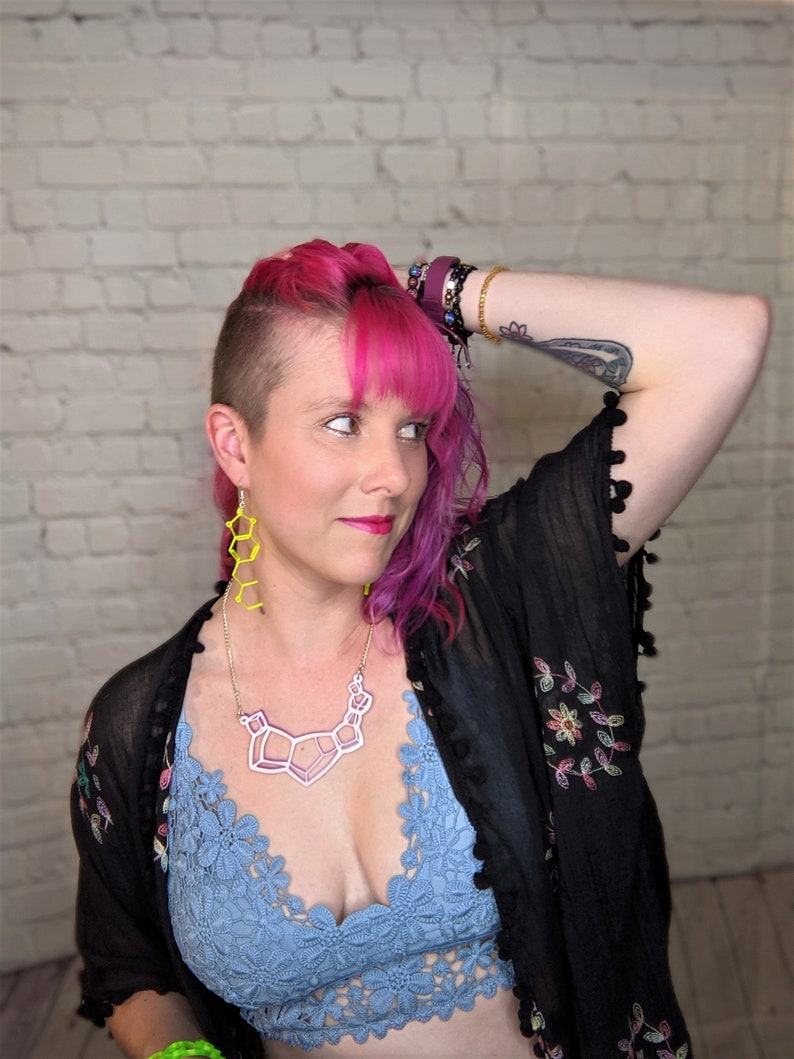 May include: A woman with pink hair and a shaved head is wearing a black sheer top with floral embroidery, a blue lace bralette, a geometric necklace, and yellow geometric earrings.