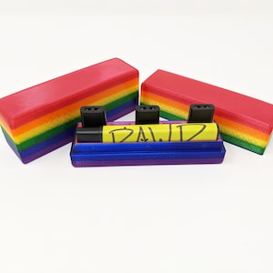 May include: A rainbow-colored storage case with a blue lid and a yellow marker with the word "DAWD" written on it. The case has three slots for small items.