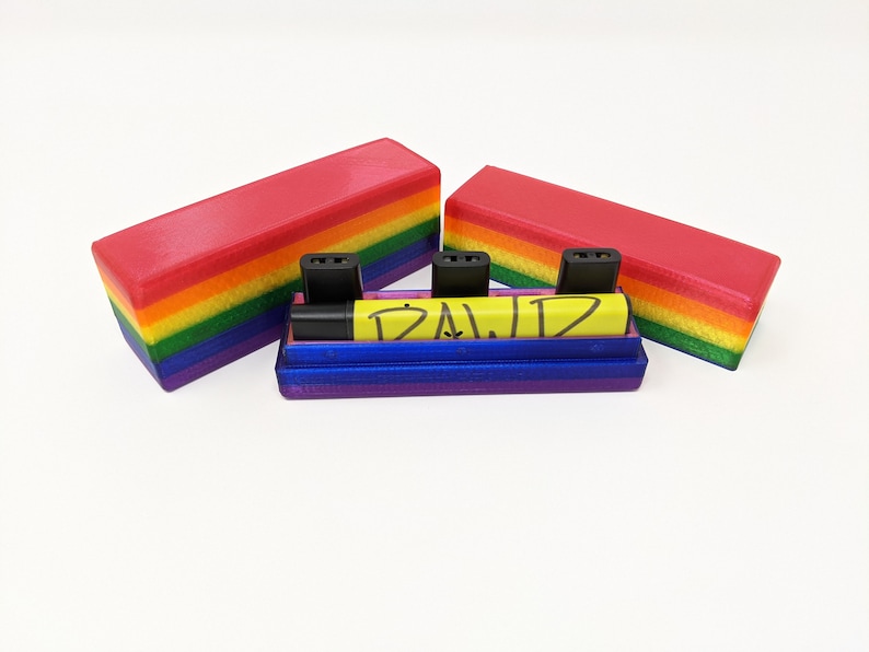 May include: A rainbow-colored storage case with a blue lid and a yellow marker with the word "DAWD" written on it. The case has three slots for small items.
