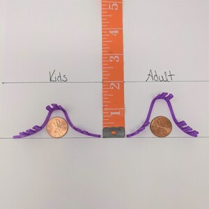 May include: Two purple plastic hair clips, one labeled "Kids" and the other "Adult", are shown next to a ruler. The ruler measures 1 to 5 inches. A penny is placed under each clip to show the size difference.