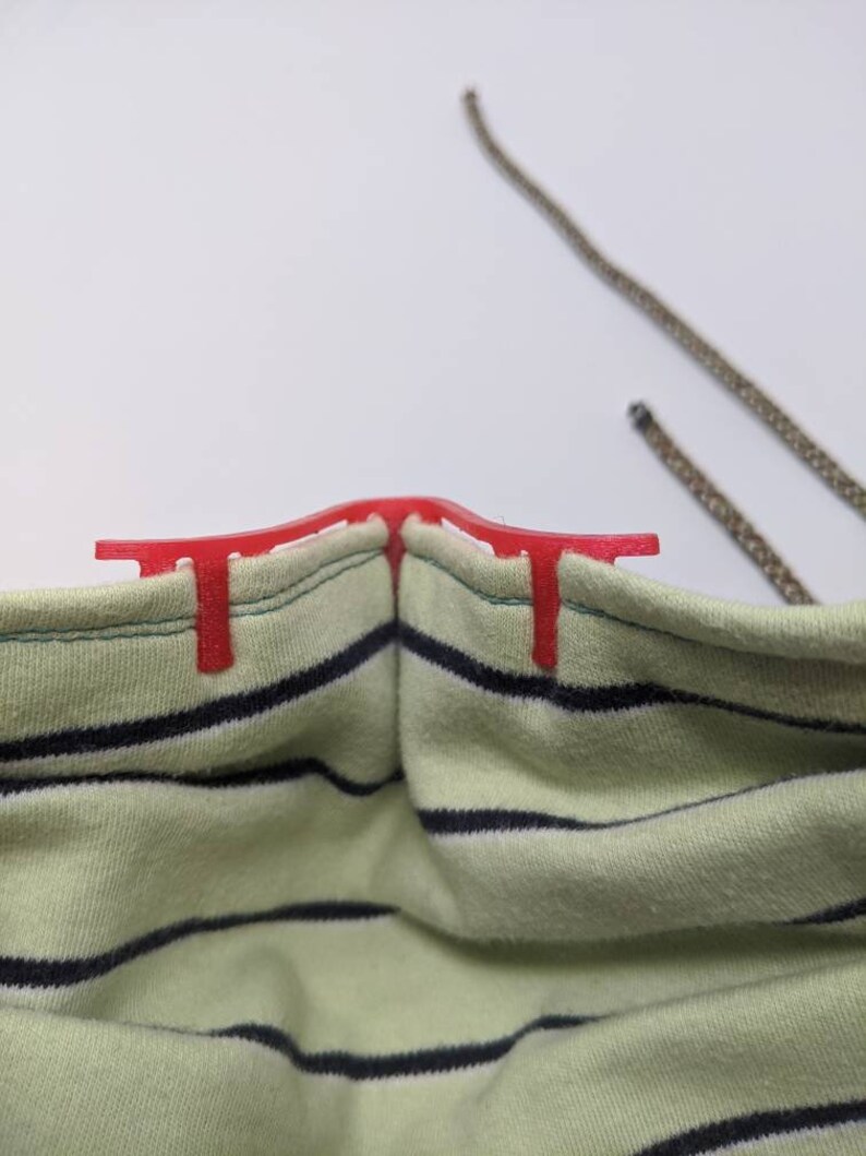 May include: A red plastic nose wire for a face mask. The nose wire is attached to a green and white striped fabric face mask.