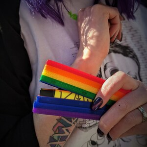 May include: A person is holding a multi-colored, rainbow-striped, plastic case with a blue and purple section. The case is open and appears to hold a small, black, rectangular object.