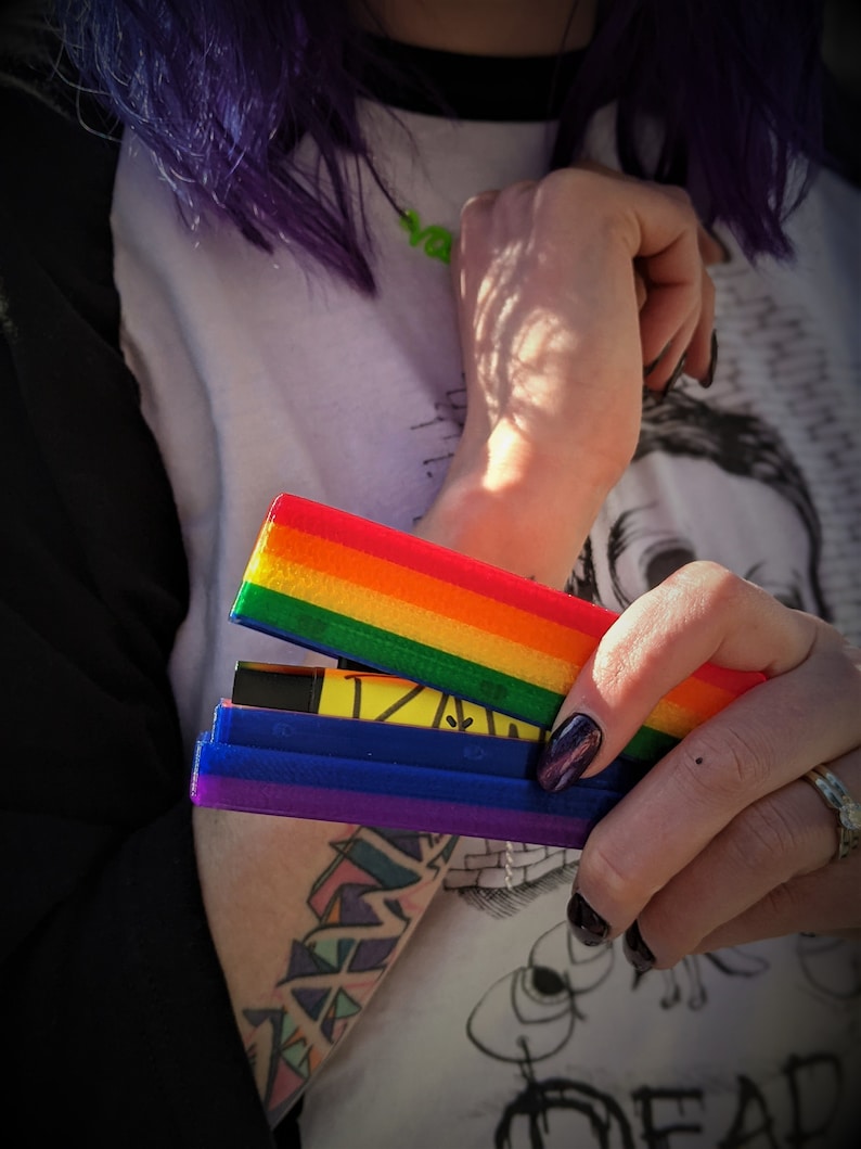 May include: A person is holding a multi-colored, rainbow-striped, plastic case with a blue and purple section. The case is open and appears to hold a small, black, rectangular object.